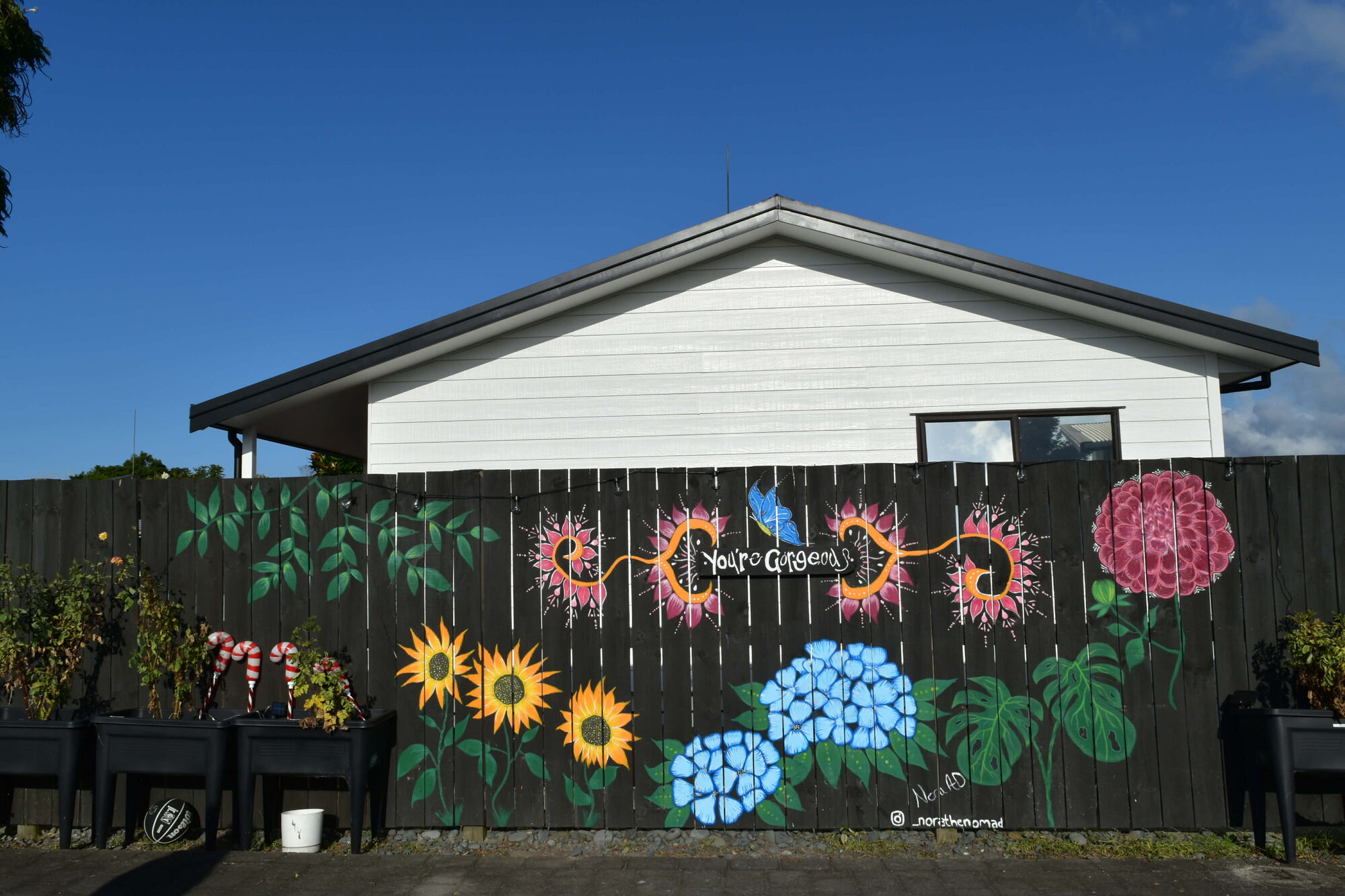 A dark fence painted with sunflowers, hydrangeas and bold blooms beside a house