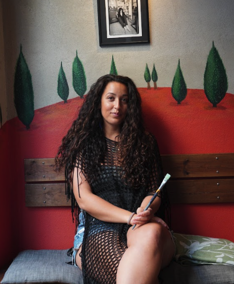 Nora seated in front of a hand-painted mural with trees and a red landscape