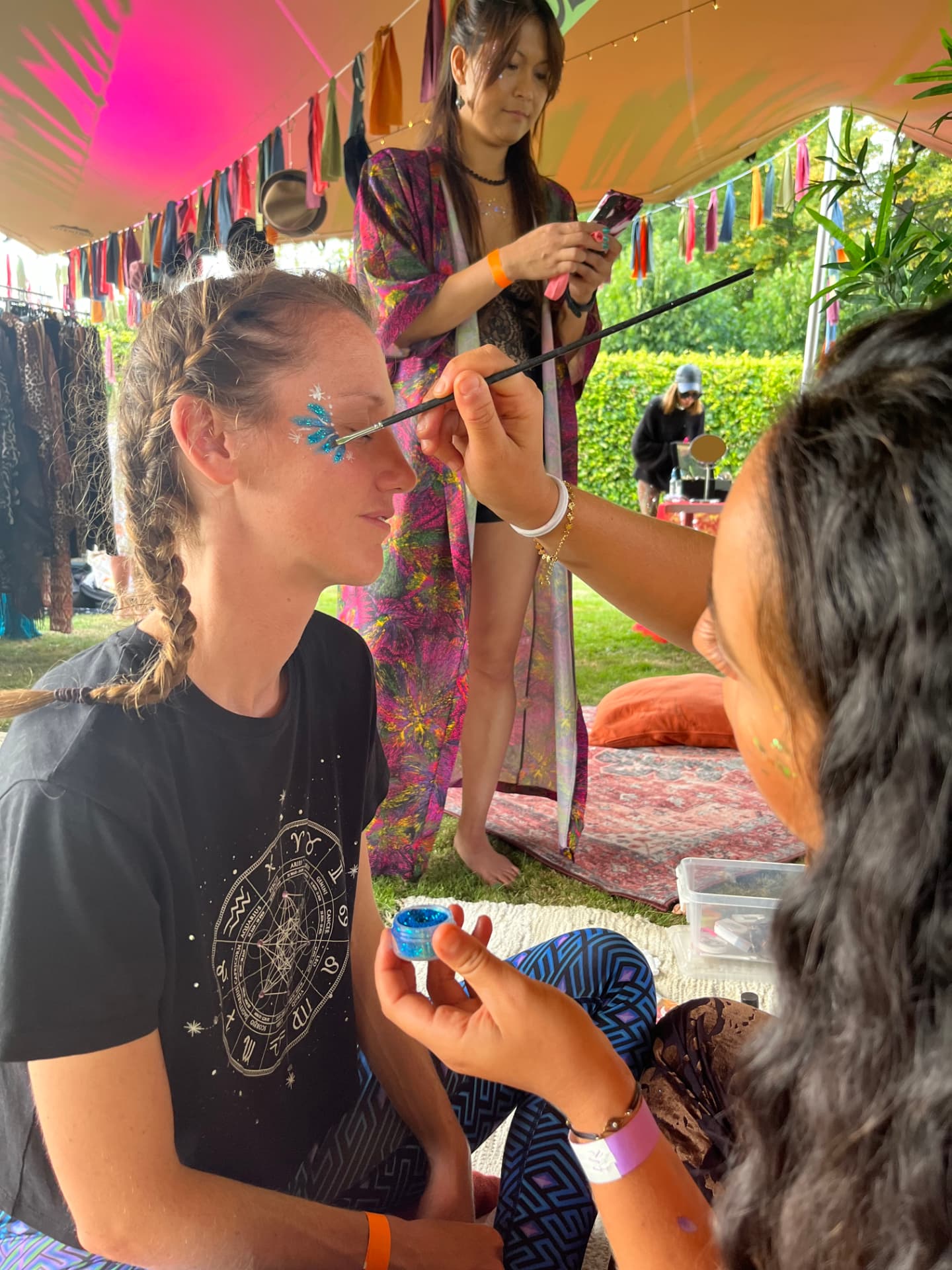 Nora applying blue glitter face paint to a festival guest under a colorful canopy
